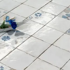 Pavimento con piastrelle quadrate, alcune decorate con fiori blu e grigi, altre bianche. Un vassoio in legno con un fiore blu e un portacandele blu completano la scena, mostrando la differenza tra mattonella e piastrella.