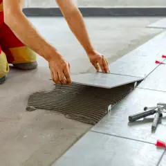 Installazione di piastrelle quadrate con stucco fresco. Un operaio in tuta rossa e gialla posa una piastrella, seguendo schemi precisi.
