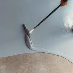 Applicazione di resina per un pavimento fai da te. Una spatola stende il materiale su una superficie grigia, mentre una persona con scarpe chiodate cammina sul cemento grezzo.