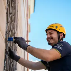 Operaio con casco giallo applica pittura su grata metallica, parte di un sistema di intonaco armato con rete in fibra di vetro.