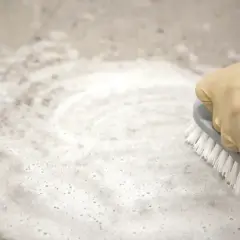 Mano con guanto giallo strofina un pavimento in gres con una spazzola, creando schiuma con ammoniaca per pulire.