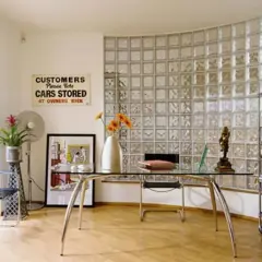 Un ufficio moderno con una parete curva in vetrocemento, scrivania in vetro, sedia, ventilatore e scaffali pieni di libri.
