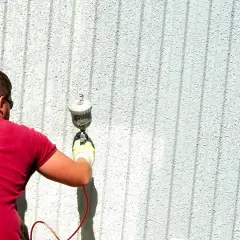 Uomo con maglietta rossa e occhiali da sole impara come verniciare a spruzzo una parete esterna con una pistola a spruzzo.