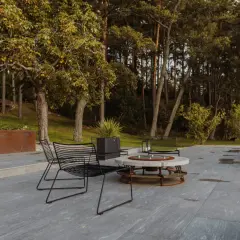 Terrazza con pavimentazione in gres flottante, sedie nere e tavolino con braciere, circondata da alberi e vegetazione.