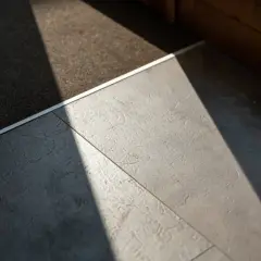 Un tappeto scuro incontra un moderno pavimento flottante grigio, illuminato da un raggio di sole diagonale.