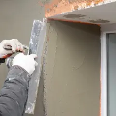 Lavoratore applica intonaco alle spallette di finestre esistenti, usando una cazzuola e rete di rinforzo.