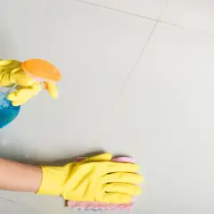 Mani con guanti gialli puliscono piastrelle dopo stuccatura con spray e panno.