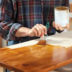 Mano che stende flatting su un piano in legno grezzo con un pennello, dando una finitura lucida e protettiva.