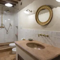 Bagno con piastrelle bianche quadrate, lavabo in cotto, specchio dorato e doccia con rubinetteria in ottone.