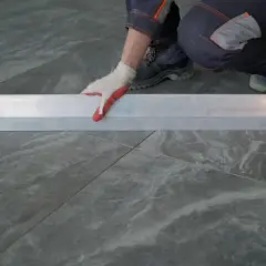 Mano con guanto bianco e rosso posiziona un profilo metallico su piastrelle grigie 60x120. Preventivo prezzo posa piastrelle 60x120.