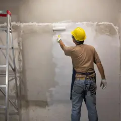 Operaio con casco giallo applica fissativo per pittura su muro grezzo con rullo, accanto a una scala metallica.