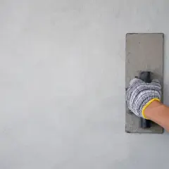 Mano con guanto che stende il rasante fino per interni su una parete grigia, creando una finitura liscia.