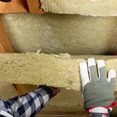 Operaio con guanti installa pannelli di lana di roccia isolante tra le travi di legno di un sottotetto.