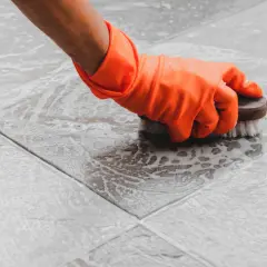 Mano con guanto arancione che lucida le piastrelle del pavimento con una spazzola, creando schiuma.