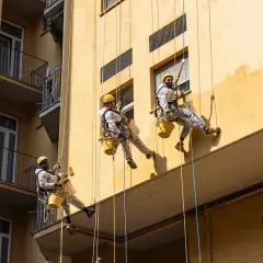 Operai in corda applicano il miglior intonaco per esterni su un edificio giallo. Lavori in quota per un risultato impeccabile.