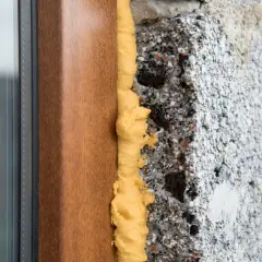 Schiume poliuretaniche espandenti sigillano il vano tra un telaio di finestra in legno e un muro grezzo, garantendo isolamento termico.