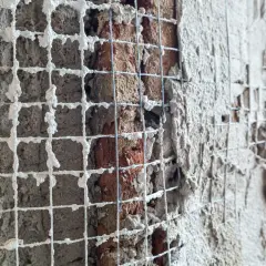 Rete metallica applicata a un muro di mattoni per un rinforzo strutturale, con intonaco fresco che la ricopre parzialmente.