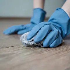Mani con guanti blu puliscono un pavimento in legno con un panno, mostrando come togliere il miele dal pavimento.