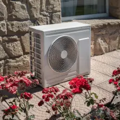 Un'unità esterna di pompe di calore, bianca e moderna, è posizionata accanto a un muro di pietra grezza, circondata da fiori rossi.