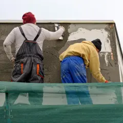 Lavoratori applicano intonaco su un muro esterno, integrando una rete per rinforzo. La rasatura del muro esterno con rete è in corso.