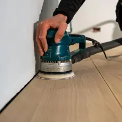 Mano che usa una levigatrice orbitale per levigare il parquet fai da te, avvicinandosi al battiscopa bianco.