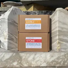 Due scatole di prodotti per la pulizia del marmo, "MarbleShine" e "Stonecare", sono posizionate tra blocchi di marmo grezzo. Ideale per sapere come lavare pavimento in marmo.