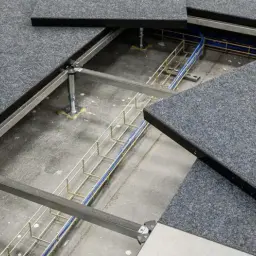 Dettaglio del pavimento galleggiante per ufficio con pannelli grigi in moquette e un'area aperta che rivela cavi e struttura metallica sottostante.