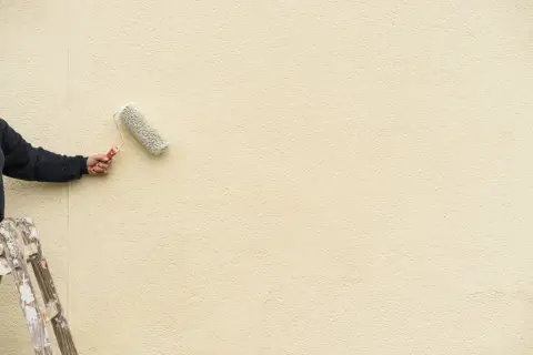 Mano con rullo per pittura facciata esterna, su scala, che rinnova il muro di colore giallo chiaro.