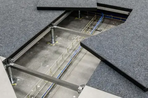 Dettaglio del pavimento galleggiante per ufficio con pannelli grigi in moquette e un'area aperta che rivela cavi e struttura metallica sottostante.