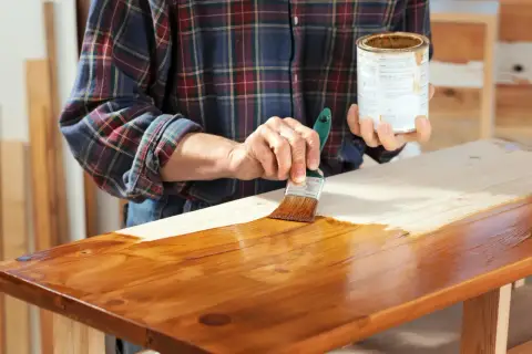 Mano che stende flatting su un piano in legno grezzo con un pennello, dando una finitura lucida e protettiva.