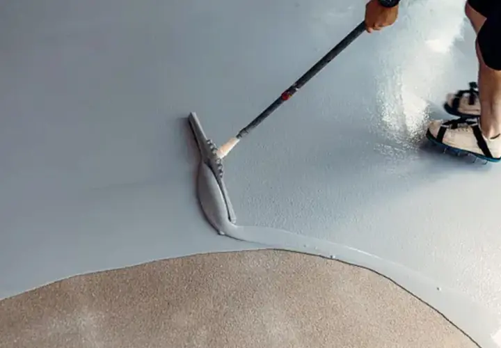 Applicazione di resina per un pavimento fai da te. Una spatola stende il materiale su una superficie grigia, mentre una persona con scarpe chiodate cammina sul cemento grezzo.