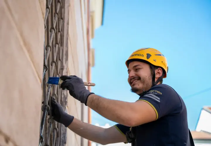 Operaio con casco giallo applica pittura su grata metallica, parte di un sistema di intonaco armato con rete in fibra di vetro.