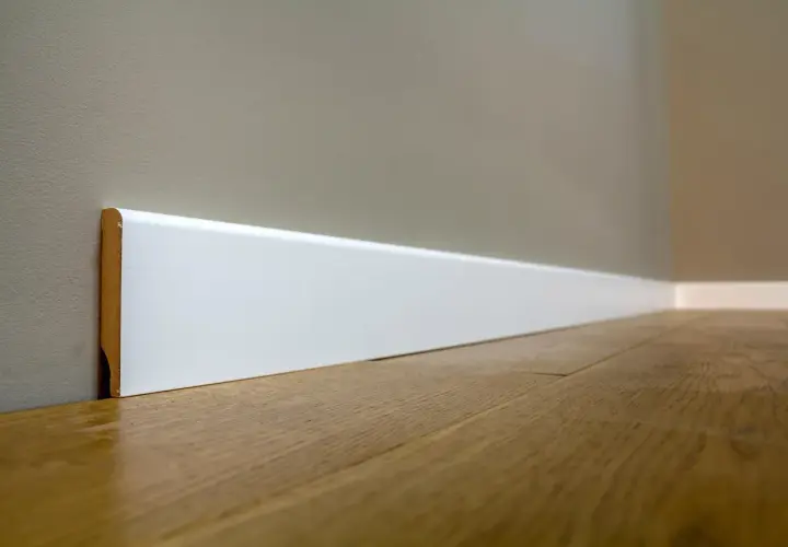 Battiscopa bianco moderno, con un'altezza che definisce lo spazio. Il legno del pavimento riflette la luce.