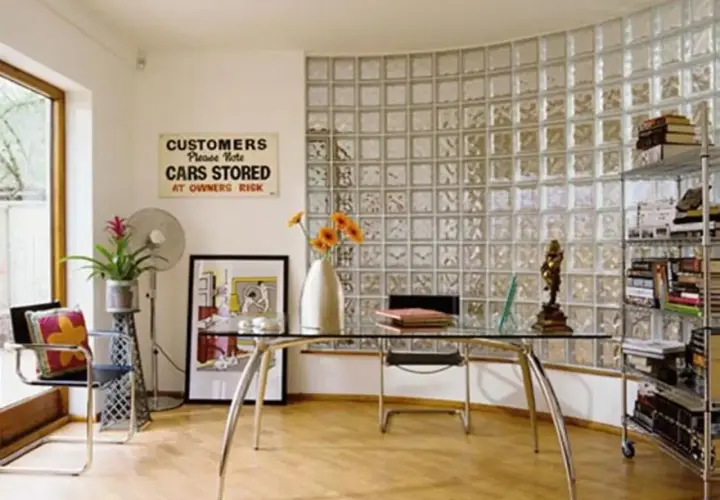 Un ufficio moderno con una parete curva in vetrocemento, scrivania in vetro, sedia, ventilatore e scaffali pieni di libri.