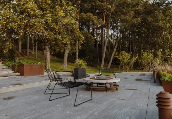 Terrazza con pavimentazione in gres flottante, sedie nere e tavolino con braciere, circondata da alberi e vegetazione.