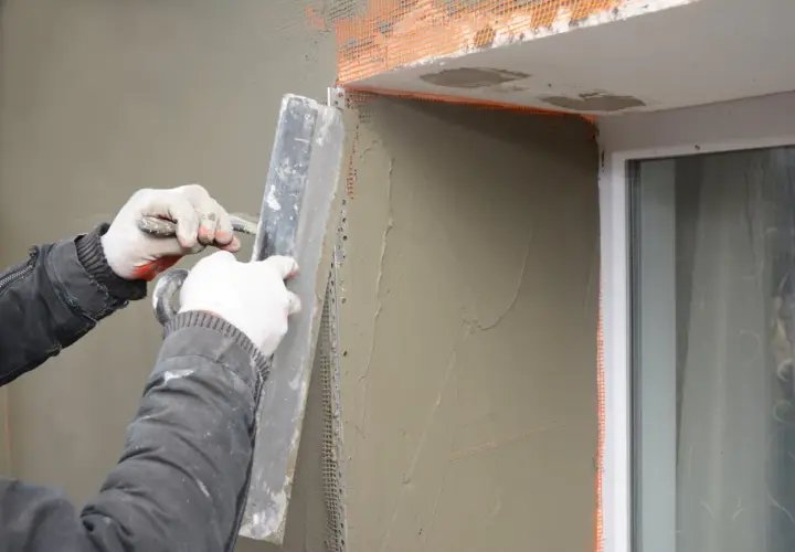 Lavoratore applica intonaco alle spallette di finestre esistenti, usando una cazzuola e rete di rinforzo.