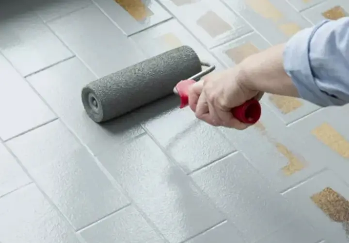 Applicazione di smalto per pavimenti: pro e contro di un restyling fai-da-te. Un rullo grigio stende uniformemente la vernice su piastrelle grigie.