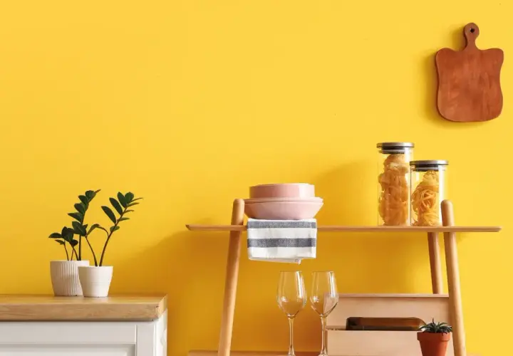 Cucina con pareti giallo vivace, dove **vernici naturali** esaltano gli arredi: piante, pasta in barattoli, piatti rosa e un tagliere in legno.