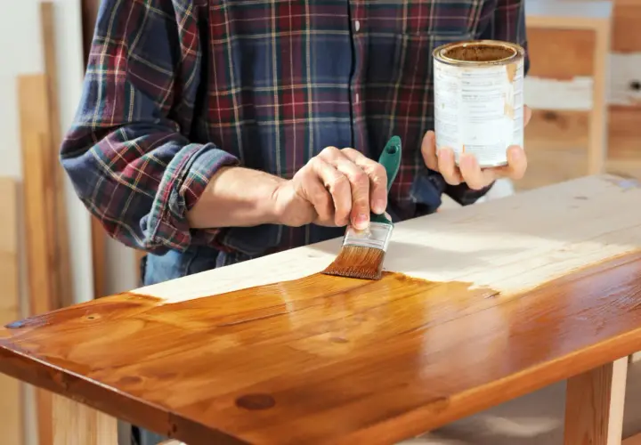 Mano che stende flatting su un piano in legno grezzo con un pennello, dando una finitura lucida e protettiva.