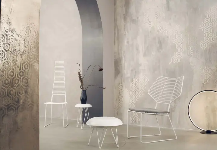 Parete con texture materica e motivi esagonali, che mostra diversi tipi di pittura decorativa. Due sedie bianche minimaliste e tavolini completano l'arredo.
