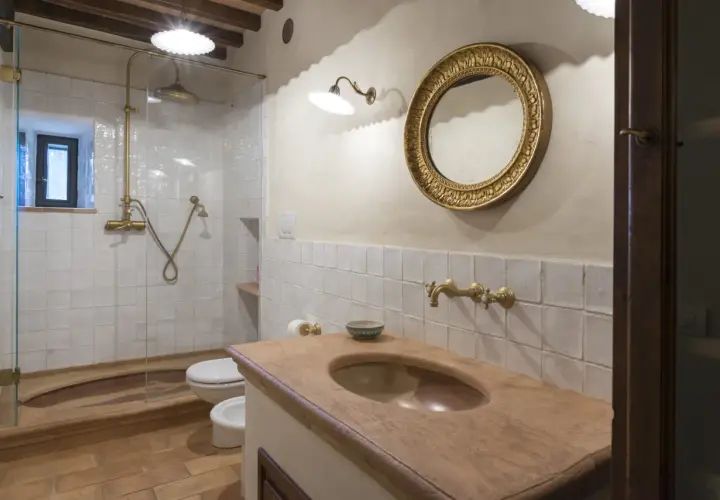 Bagno con piastrelle bianche quadrate, lavabo in cotto, specchio dorato e doccia con rubinetteria in ottone.