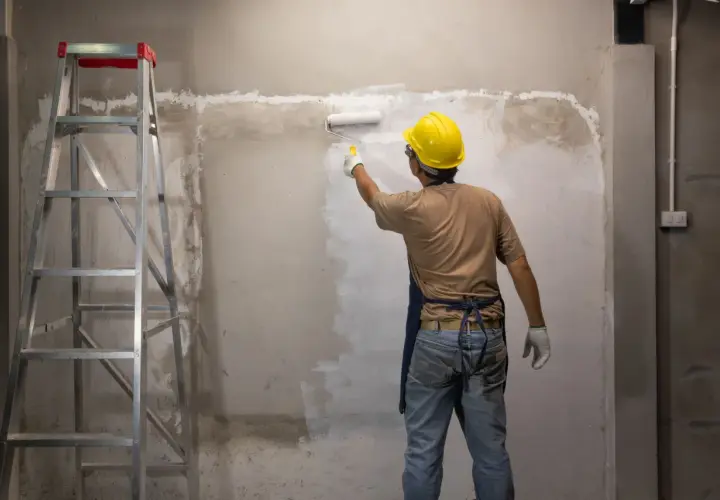 Operaio con casco giallo applica fissativo per pittura su muro grezzo con rullo, accanto a una scala metallica.