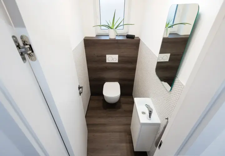Bagno piccolo stretto con WC, lavandino bianco, specchio e pianta. Pareti con piastrelle bianche a mosaico e inserto in legno scuro.