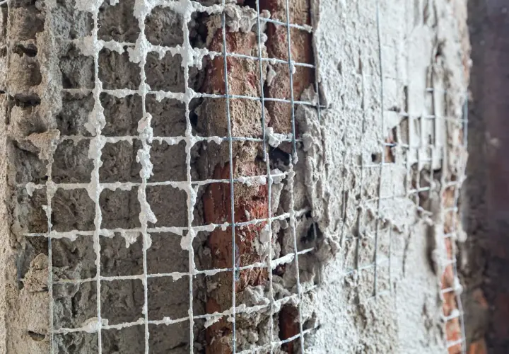 Rete metallica applicata a un muro di mattoni per un rinforzo strutturale, con intonaco fresco che la ricopre parzialmente.