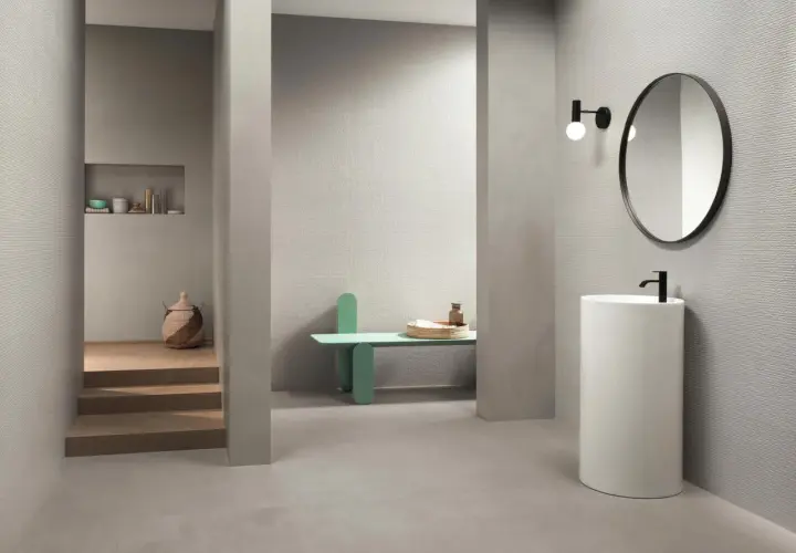 Bagno moderno con rivestimento uguale al pavimento, lavabo a colonna, specchio rotondo e nicchia con oggetti decorativi.