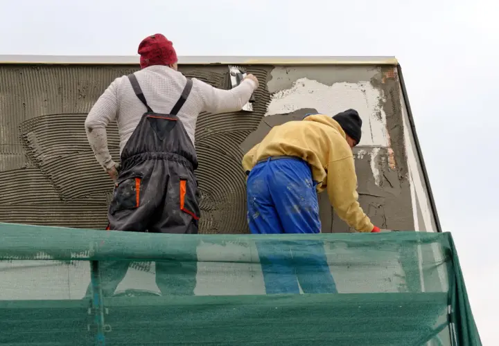 Lavoratori applicano intonaco su un muro esterno, integrando una rete per rinforzo. La rasatura del muro esterno con rete è in corso.