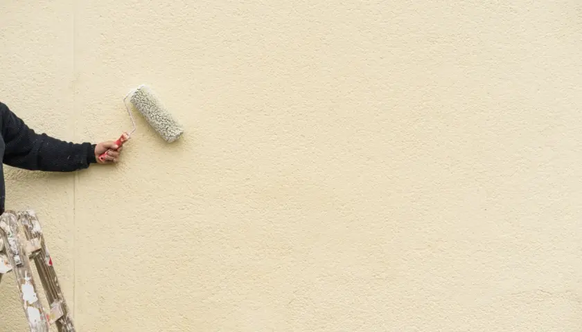Mano con rullo per pittura facciata esterna, su scala, che rinnova il muro di colore giallo chiaro.