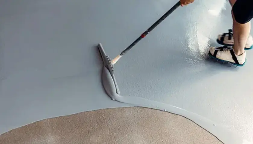Applicazione di resina per un pavimento fai da te. Una spatola stende il materiale su una superficie grigia, mentre una persona con scarpe chiodate cammina sul cemento grezzo.