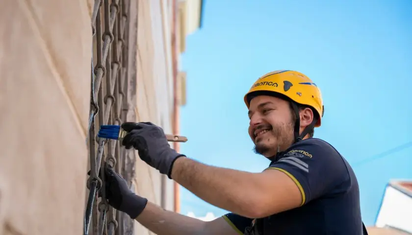 Operaio con casco giallo applica pittura su grata metallica, parte di un sistema di intonaco armato con rete in fibra di vetro.