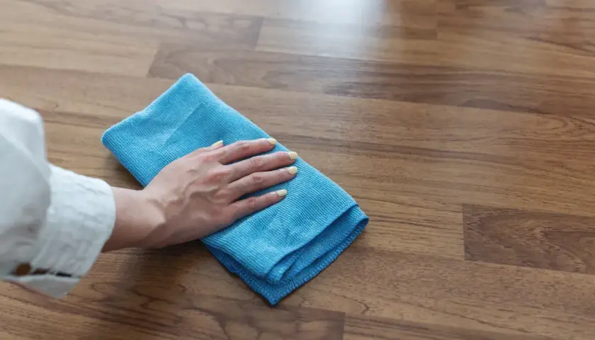 Mano con panno blu che pulisce un pavimento in legno, mostrando come togliere la colla dal legno con delicatezza.